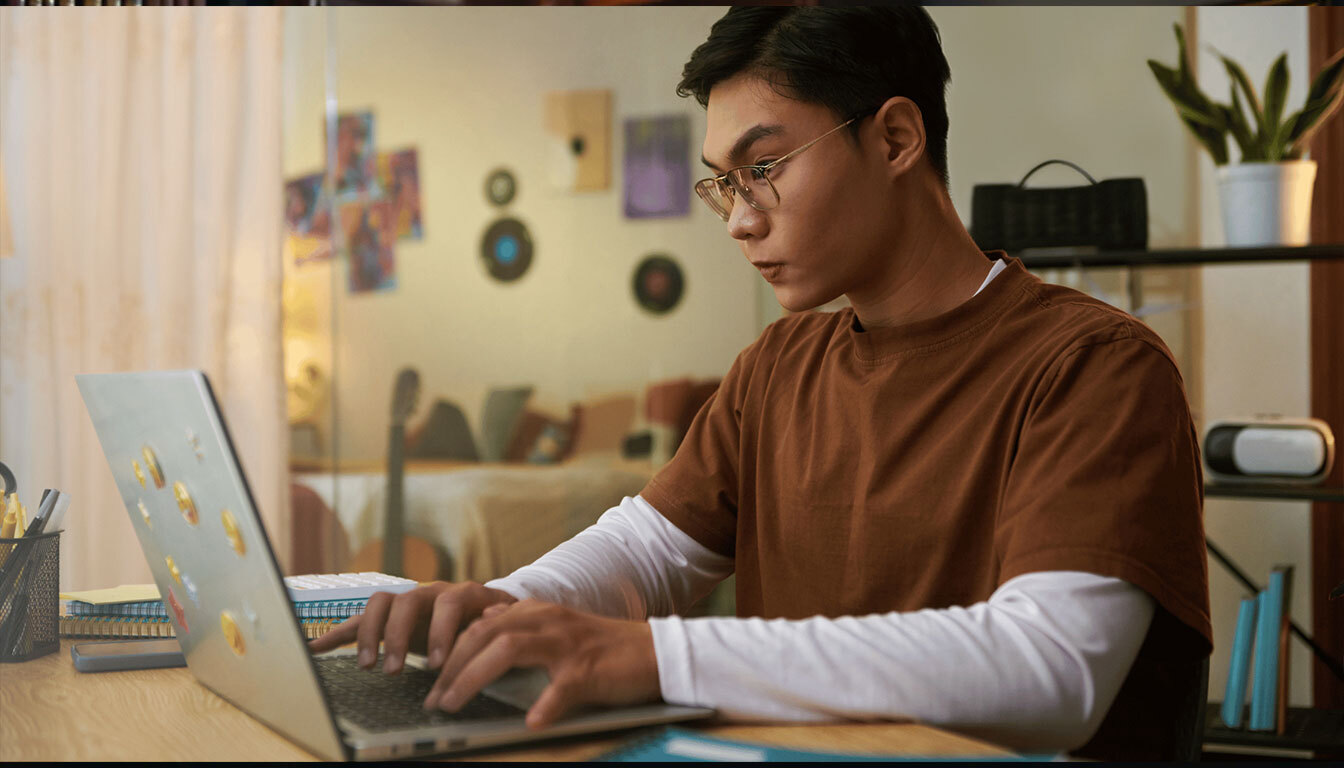 A man wearing glasses focuses intently on his laptop while working in a cozy, well-lit environment